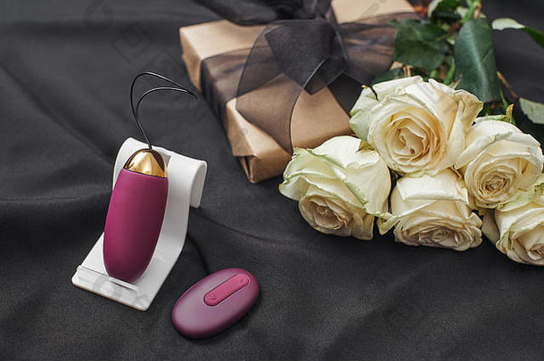 情人节礼物创意。紫色振动蛋的特写镜头，远处美丽的白玫瑰点缀在黑色丝绸上。玩具。成人礼物