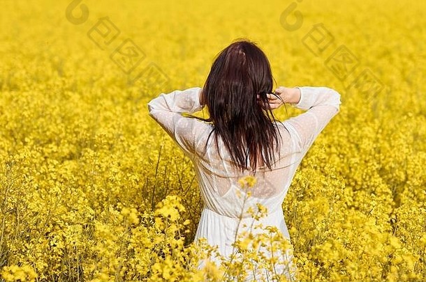 身穿白色连衣裙的年轻女子站在油菜地里。开放的黄色花朵与空间