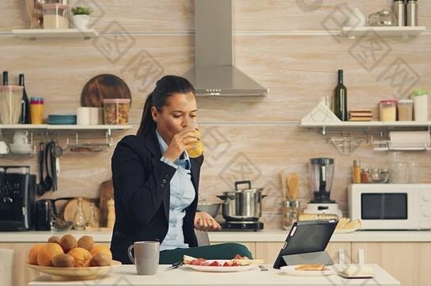 女商人在早餐时喝新鲜果汁，然后在工作前观看平板电脑上的视频。女商人上班前在网上阅读最新新闻，在厨房里使用现代技术，同时吃健康的一餐