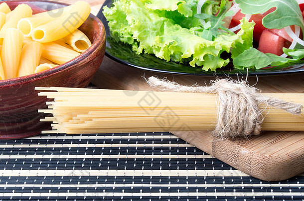 桌子的细节和健康食品的特写-生菜在一个黑色的盘子里，意大利面在一个木制的碗里和一堆意大利面在一个木制的板上
