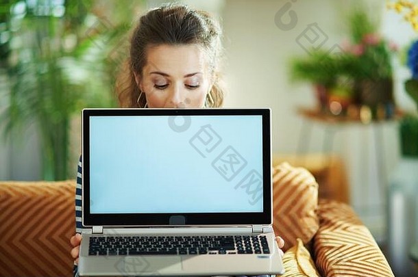 时尚的中间年龄家庭主妇蓝色的上衣条纹夹克现代生活房间阳光明媚的一天坐着沙发上显示移动PC空白屏幕