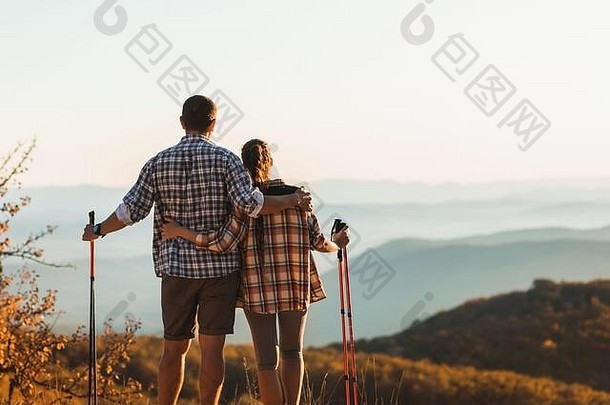 一对夫妇带着<strong>登山杖</strong>在秋天的山上徒步旅行。北欧人走在户外，从后面看。旅游生活方式。