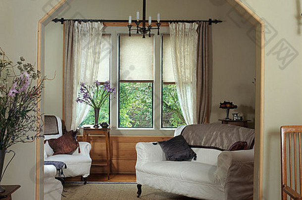 从门口可以看到沙发上的白色宽松被子和小客厅延伸部分的椅子，带有白色薄纱和米色窗帘