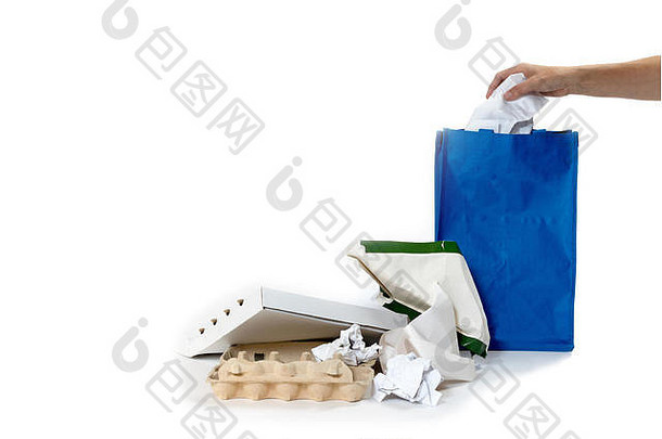 一堆废纸躺在白色的背景上。女人的手正在把用过的纸塞进一个空的蓝色袋子里。