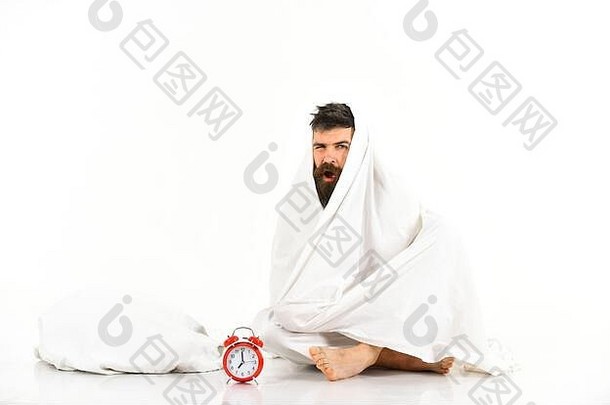 瞌睡虫概念。那个张脸平静、打呵欠的男人坐在闹钟旁边的毯子下。男人想躺在床上，白色背景。留胡子的家伙躲在毯子下面。