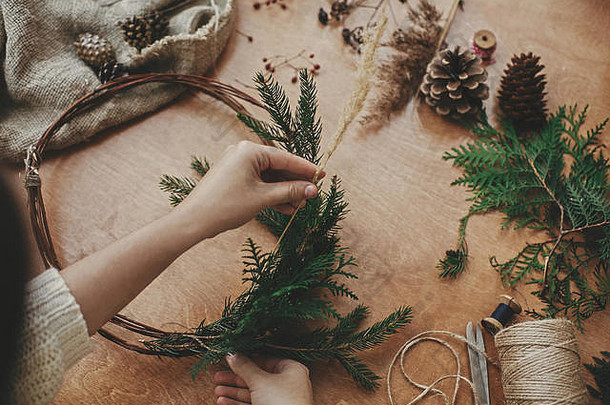 圣诞花圈工作坊。手拿着药草、冷杉枝、松果、浆果、线、剪刀放在木桌上。制作质朴的圣诞花环。A.