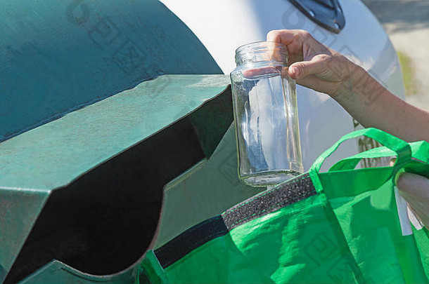 该名女子正将一个玻璃瓶从绿色便携袋扔到用于分类玻璃生活垃圾的绿色容器中。