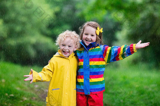 小男孩和小女孩在多雨的夏季公园里玩耍。穿着五颜六色彩虹夹克和防水靴的孩子们跳进了水坑