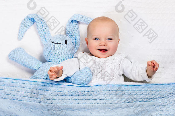 有趣的婴儿穿温暖的针织夹克玩玩具兔子放松白色电缆针织毯子阳光明媚的托儿所
