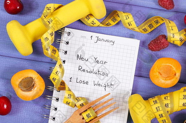 写在笔记本上的新年决心或减肥目标、新鲜水果、健身哑铃和木叉卷尺