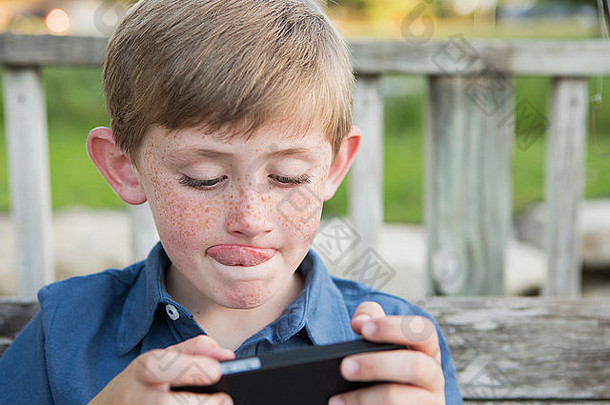 一个小男孩使用手持电子平板电脑或游戏，全神贯注，伸出舌头。