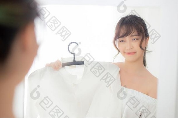 美丽的亚洲年轻女子微笑着穿着衣服，在房间里用现代的反光镜试穿衣服，女孩选择衣服