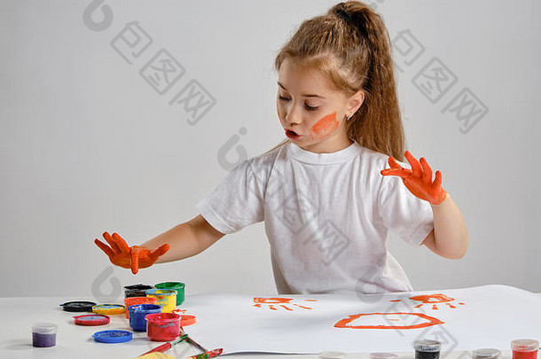 穿着白色t恤的小女孩坐在桌旁，手里拿着<strong>惠</strong>特曼和五颜六色的颜料，用手在上面画画。白色的隔离带。中等特写镜头。