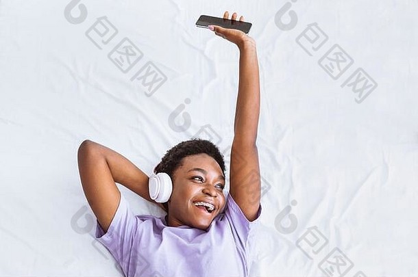 欣赏喜爱的音乐。女孩躺在床上，带着智能手机，戴着耳机<strong>听歌</strong>曲