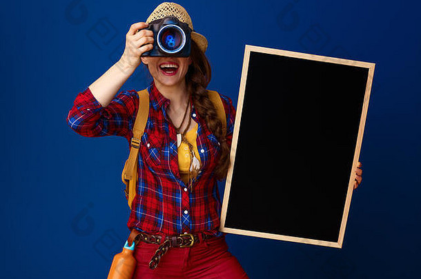 寻找鼓舞人心的地方。带着背包和黑板的年轻女游客用<strong>蓝色</strong>背景上隔离的单反相机拍照