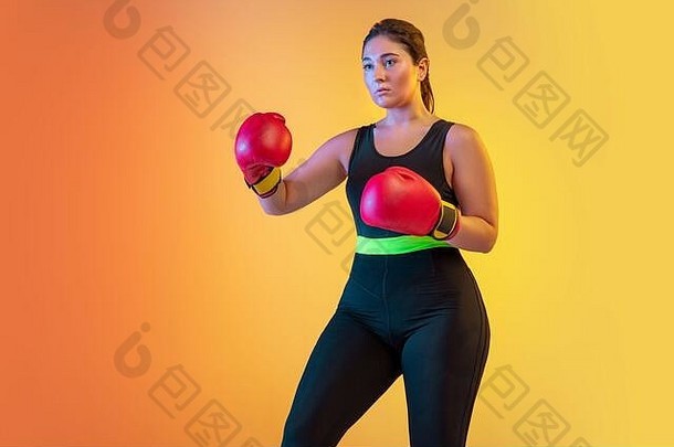 年轻的白人加大体型女模特在霓虹灯下的渐变橙色桃色背景上训练。戴拳击手套训练。体育观念、健康生活方式、身体积极、平等。