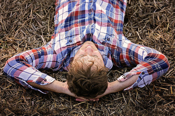 秋天的一天，一个穿着法兰绒衬衫的年轻农民正在干草捆上休息和放松。