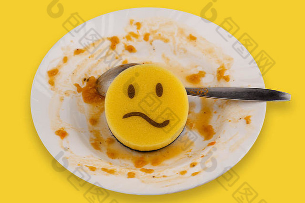 洗碗概念黄色海绵，在白色盘子和勺子上放着食物残渣，脸上带着悲伤的表情