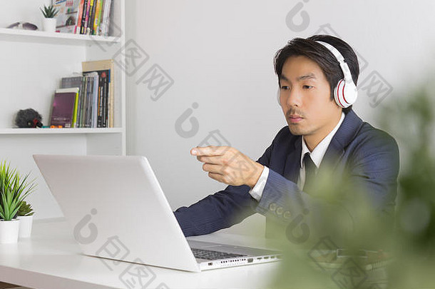 亚洲男士呼叫中心穿着西装，戴着耳机，认真回答客户的问题。亚洲男子呼叫中心在笔记本电脑前工作