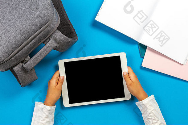 浅蓝色背景上的平板电脑、背包和彩色纸笔记本的儿童手俯视图