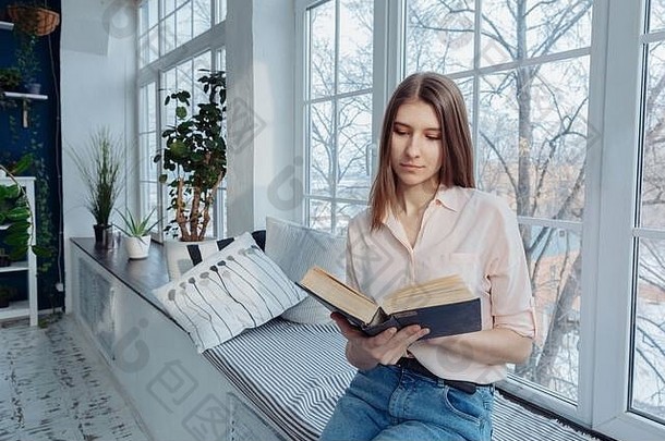 一个女孩正在窗边看书。室内植物在背景中可见