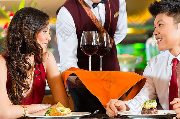 在高级餐厅或酒店的托盘上，亚洲华人侍者或服务员为男女或夫妇提供盛在玻璃杯中的红酒