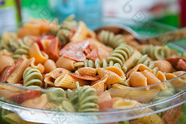 在一个透明的玻璃碗里放着一份色彩斑斓的夏季<strong>意大利面</strong>沙拉，里<strong>面</strong>有蔬菜<strong>意大利面</strong>和<strong>意大利</strong>辣香肠。