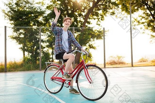 一个快乐的男孩，金发，穿着短裤和休闲衬衫，在篮球场上骑着自行车挥手打招呼。快乐的男孩