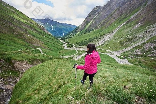 背视图：年轻女子手持<strong>登山杖</strong>独自在山中徒步旅行。女旅行者在青草丛生的山谷里走下山。旅游、旅游和徒步旅行的概念。