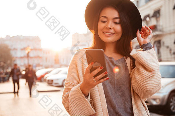 在城市街道上，一位年轻美丽、面带微笑的亚洲女子一边触摸帽子一边用智能手机查看新闻