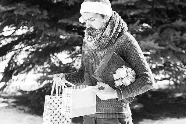 戴着圣诞老人帽子的惊讶男人，留着胡子的时髦男人，胡子上覆盖着白霜，在自然背景下，在阳光明媚的冬日打开礼品袋作为礼物