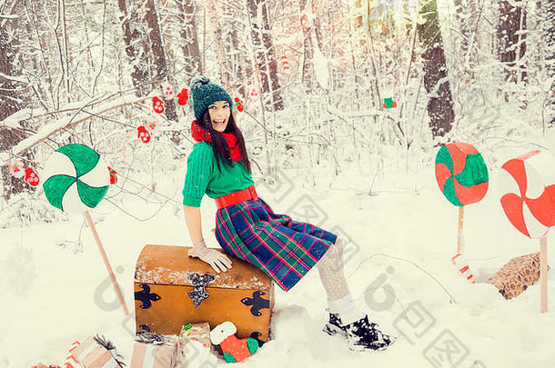 在雪地下的冬季森林里，一个黑发女孩戴着绿色帽子，穿着精灵圣诞老人的助手们传统的花朵服装，手里拿着一箱礼物和巨大的糖果