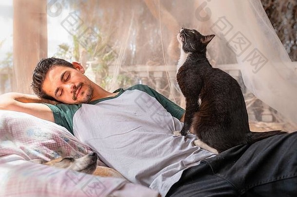 这个年轻人睡在花园里的扶手椅上，睡在蚊帐下。一只猫坐在男人的一边