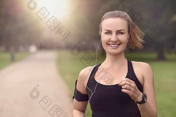 一位漂亮的运动型女子戴着耳机在公园慢跑，对着相机微笑，半身照，并留有复印空间