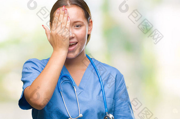 年轻的白人女医生穿着隔离背景的医疗制服，用手捂住一只眼睛，脸上带着自信的微笑，脸上带着惊喜的表情