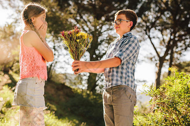 一个男孩站在公园里用鲜花向他的女朋友求婚的侧视图。男孩给一个站在公园里的女孩送花