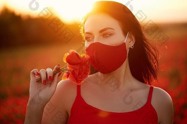 时尚隔离外观。一位年轻女子戴着红色抗病毒面具，背景是一片红花地里的夕阳。夏季时装、模特