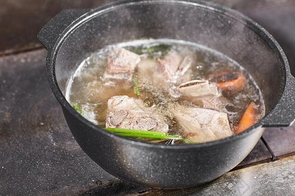 传统的牛肉汤和蔬菜，骨头和配料在锅里，烹饪食谱。用勺子在深色石头背景上煮汤。顶部