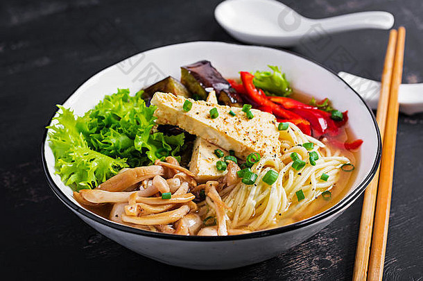 亚洲纯素面条汤，配豆腐奶酪、石磨菇和油炸茄子，背景为深色。