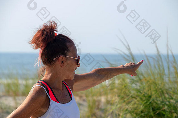 上身体视图成熟的婴儿潮高加索人女人穿运动服装太阳镜站海滩沙子沙丘高草点
