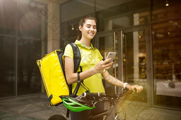 一位身穿黄色衬衫的年轻女子用小玩意跟踪城市街道上的秩序，运送食物。使用在线应用程序接收和跟踪发货地址的快递员。现代技术。