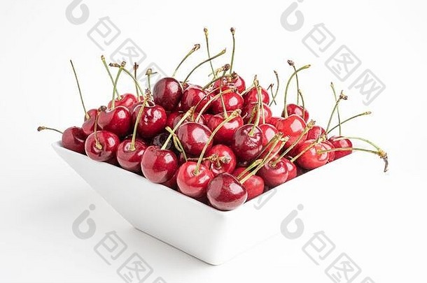 一束新鲜的红樱桃排列在一个方形的瓷碗里，背景是纯白的。