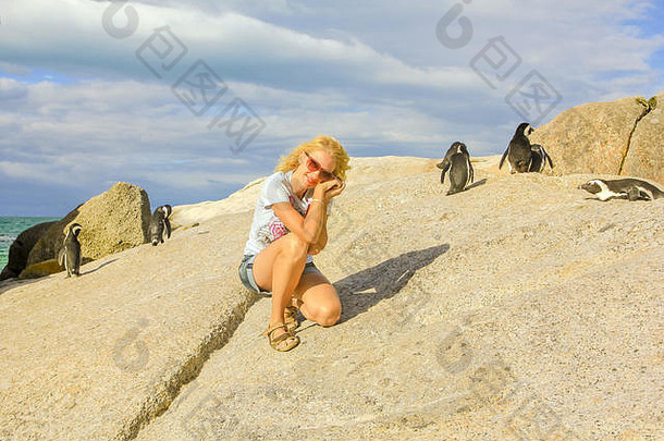 夏天，一位快乐的金发白种女人站在博尔德海滩的岩石上。博尔德海滩是一群非洲企鹅的热门旅游目的地。南非西开普省开普敦附近的开普敦半岛。