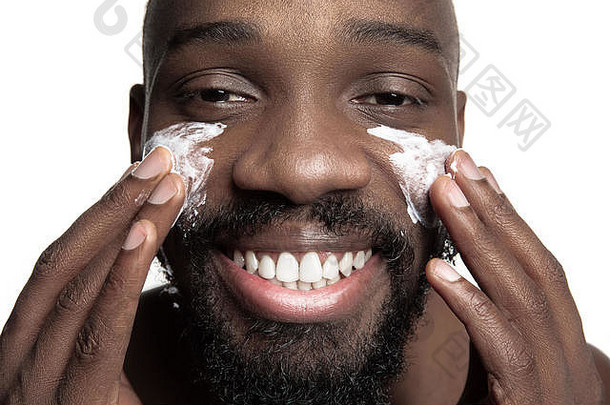 年轻的非裔美国人在白色背景下用<strong>眼霜</strong>。摄影棚里一个年轻快乐微笑的非洲男人的肖像。高级时尚男模特。