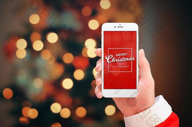 白色手机在圣诞老人的手上特写。手机屏幕上显示圣诞快乐。背景是圣诞彩灯。