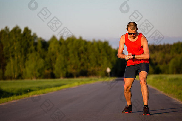 夏日夕阳中在乡村公路上跑步的男子。检查<strong>运动</strong>手表，准备跑步。<strong>青年运动</strong>员男子训练与实践