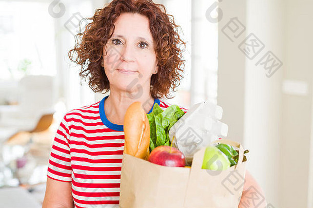 一位年长的女士拿着装满超市新鲜食品的纸袋，脸上带着自信的表情，表情聪明，思想严肃