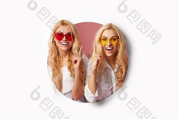 两个戴着心形太阳镜的年轻快乐、震惊的女人的肖像，透过<strong>墙</strong>上的白色孔洞望去。大减价。滑稽的面孔。空位