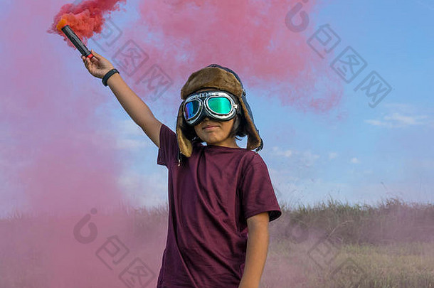 戴着头盔和飞机谷歌的小男孩站在绿色的田野上，冒着五颜六色的烟雾，假装是飞行员
