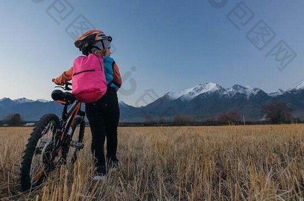一个白人孩子在麦田里骑自行车。小女孩在美丽的雪山背景下走着<strong>黑橙</strong>色的自行车。